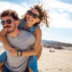 Junger Mann mit Sonnenbrille nimmt junge Frau mit Sonnenbrille am Stand huckepack